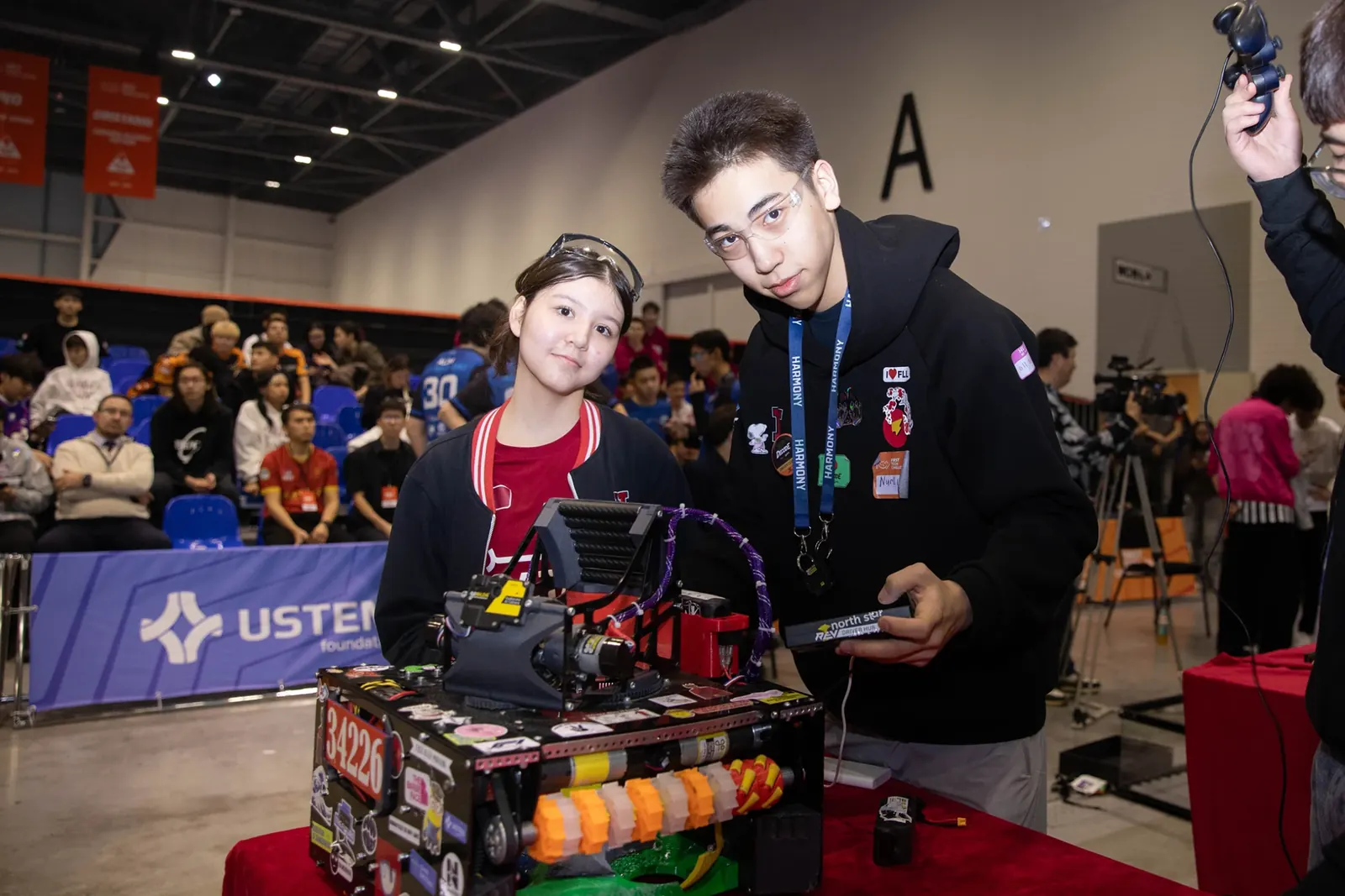 Қыз бен жігіт 34226 нөмірлі FIRST Tech Challenge роботымен, артында көрермендер мен USTEM баннері